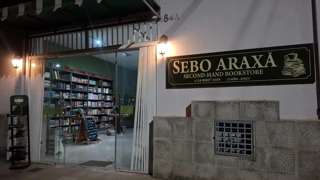 Fachada do Sebo Araxá à noite: letreiro iluminado sobre a entrada aberta, com estantes de livros visíveis lá dentro.
