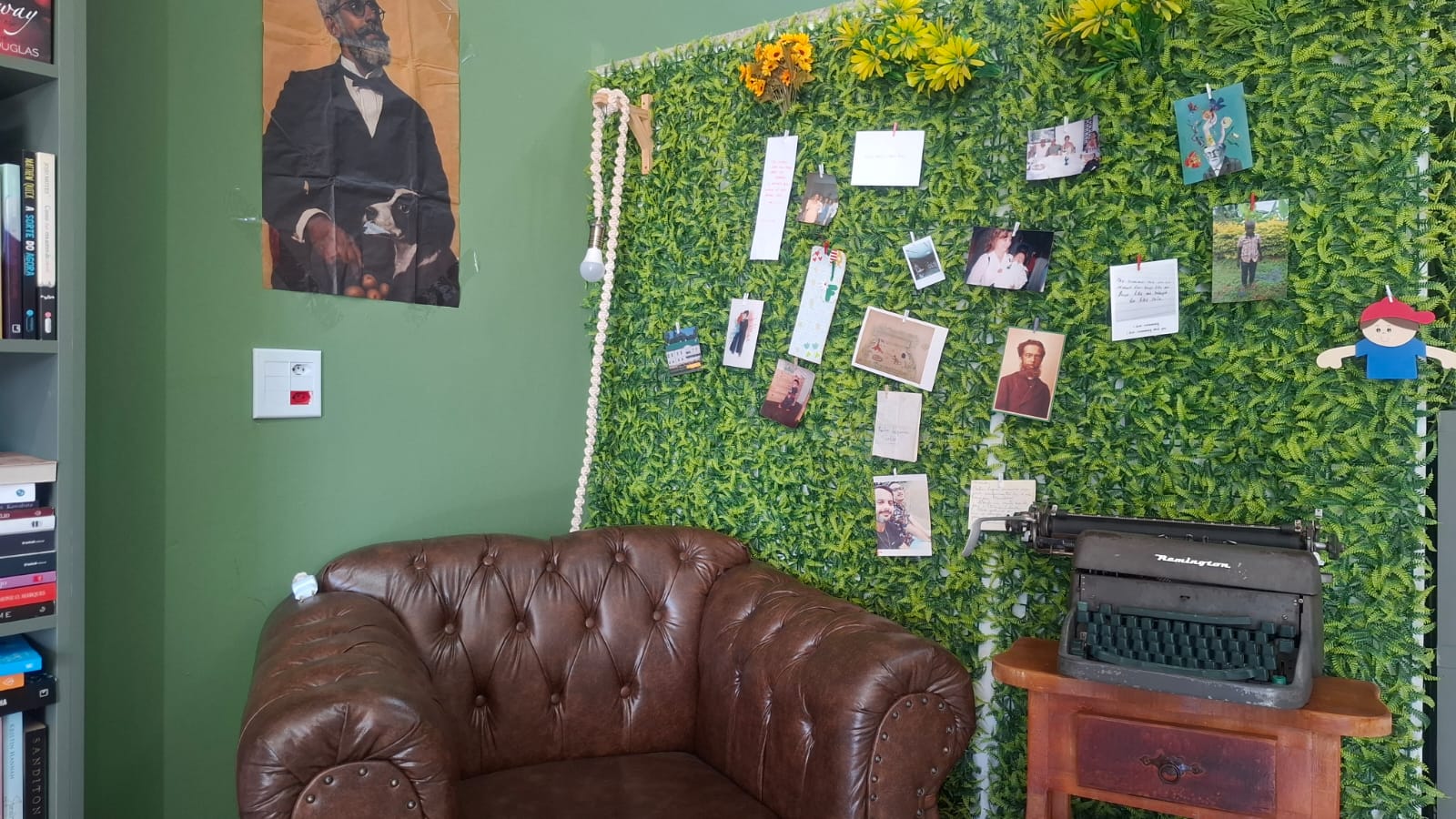 Poltrona de couro do Sebo Araxá num canto de leitura, com retrato antigo, máquina de escrever e mural verde coberto de fotos presas por pregadores.