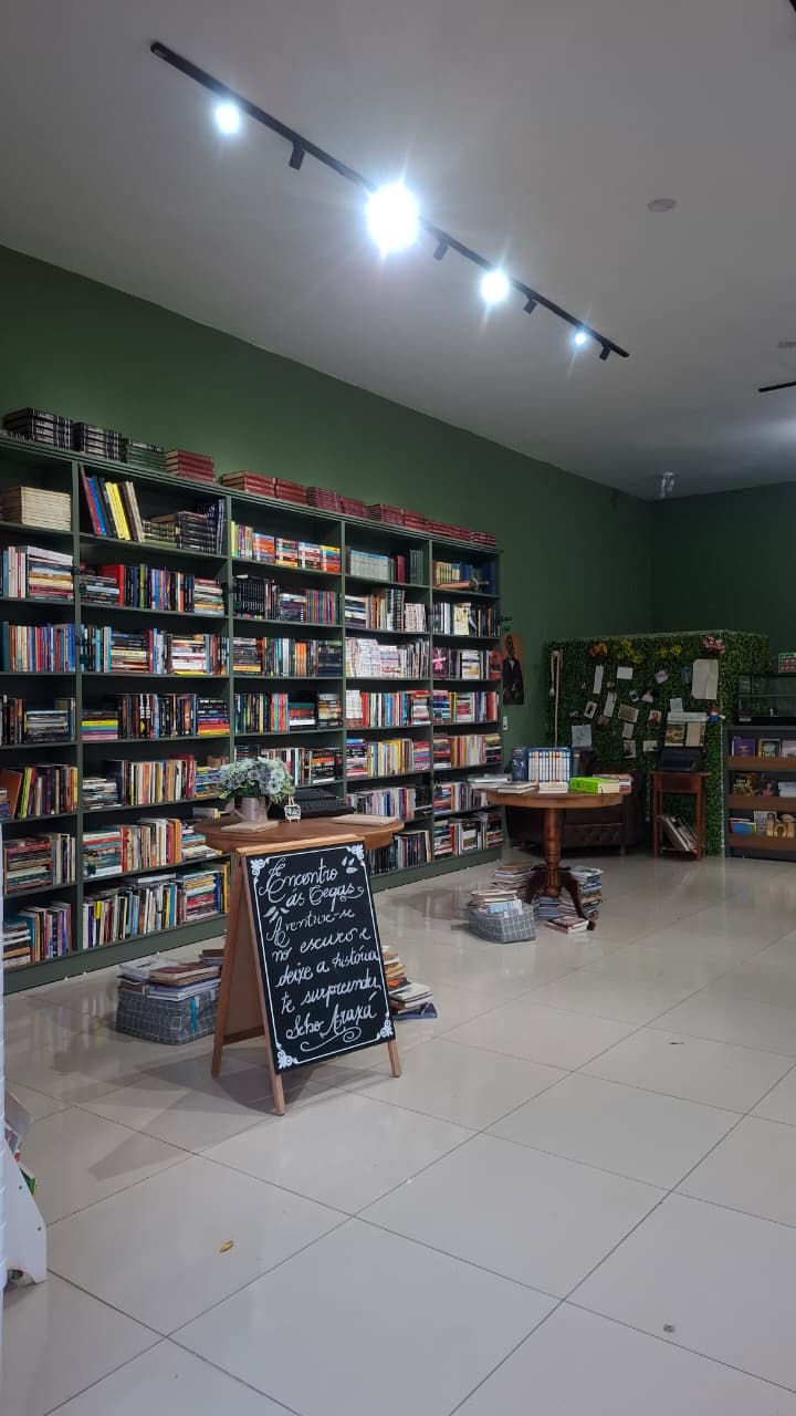 Salão do Sebo Araxá visto da entrada: estantes verdes cheias de livros, mesa com quadro-negro de recados e luminárias de trilho no teto.